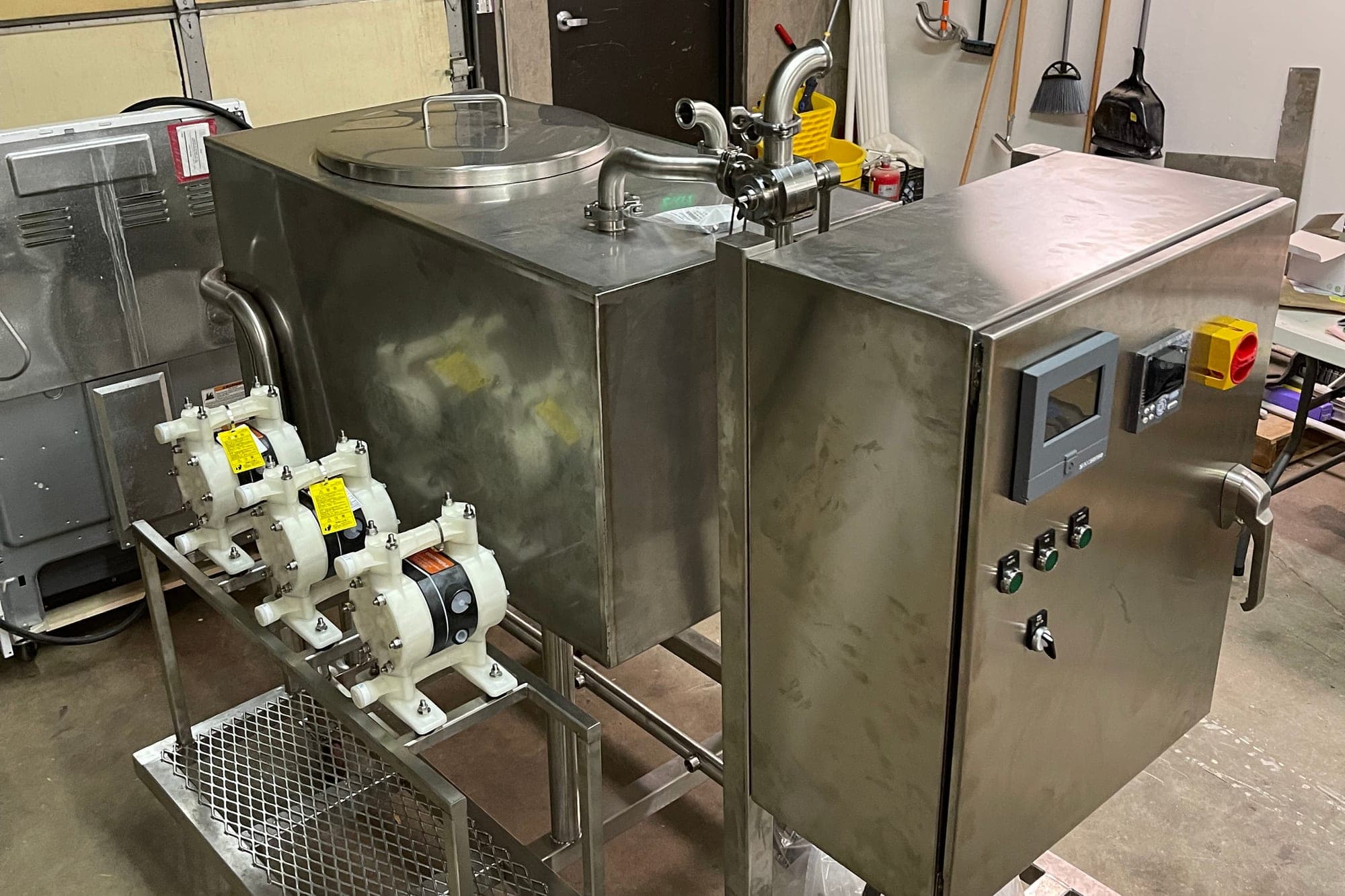 A completed stainless process skid in the Launch Partners shop — jacketed tank, three diaphragm pumps on a rail, and a stainless control cabinet with HMI and E-stop